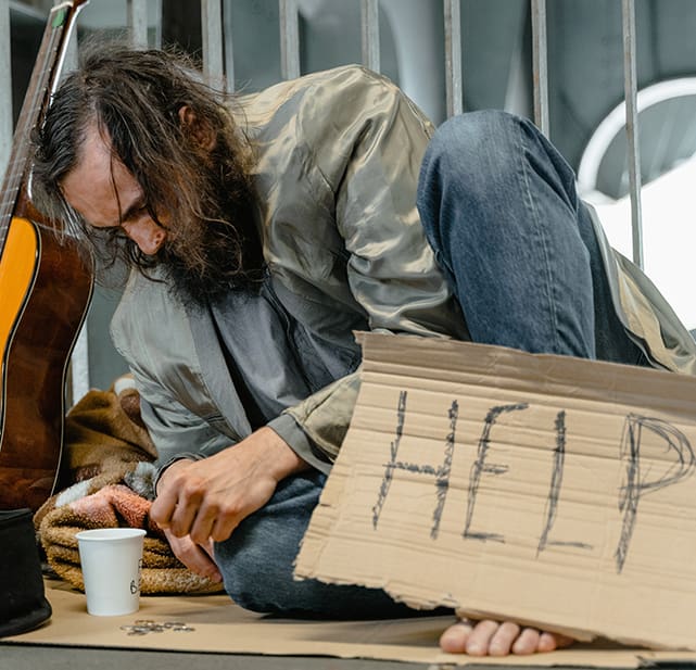 A man sitting on the ground writing help on cardboard.